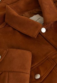 Chaqueta de ante marrón con una textura suave, con un cuello rígido, bolsillos con solapa y cierres de botón a presión blancos. Forro interior visible.