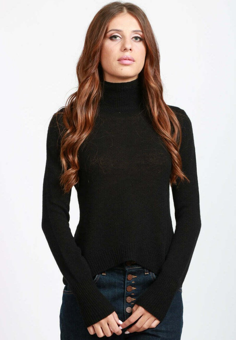Giovane donna con lunghi capelli castani ondulati, che indossa un maglione a collo alto nero e jeans blu scuro, in piedi contro uno sfondo bianco.