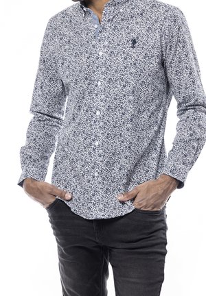 Mann trägt ein langärmeliges weißes Hemd mit marineblauem Blumenmuster und schwarze Hose, steht mit den Händen in den Taschen.