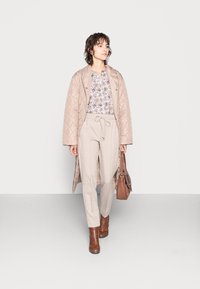 Casaco longo acolchoado bege claro, blusa com padrão floral, calças cinza claro com cordão e botins castanhos. A portar uma mala castanha.