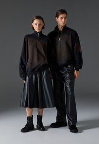 Felpe con zip nere con effetto sfumato, abbinate a una gonna in pelle nera e pantaloni. Entrambi indossano stivali militari neri, su uno sfondo grigio.