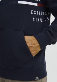 Tengerészkék pulóver elülső zsebbel, fehér felirattal és színes vízszintes csíkokkal a mellkason; puha, struktúrált anyag.