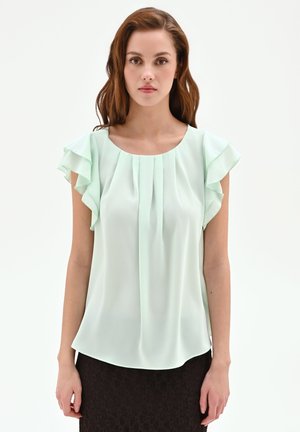 Giovane donna che indossa una blusa verde chiaro senza maniche con spalle arricciate e una gonna in pizzo nera, in piedi davanti a uno sfondo bianco.