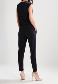 Svart, ärmlös jumpsuit med avslappnad passform, elastisk midja och smalnande ben. Bärs med beiga högklackade skor, mjuk tygtextur.