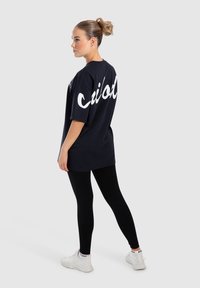 Černé oversized tričko s bílým zakřiveným nápisem na zádech, v kombinaci s černými legínami a bílými teniskami. Hladká látka, příjemný střih.