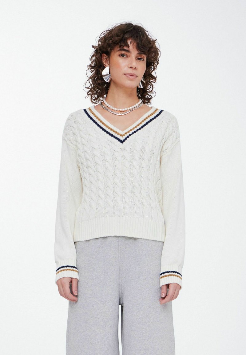 White cable-knit sweater with V-neck, featuring navy and gold stripes on the neck and cuffs, paired with gray sweatpants.