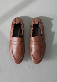 Scarpe slip-on in pelle marrone con punta arrotondata, apertura elasticizzata e dettaglio di cucitura decorativa sulla parte superiore. Texture liscia.