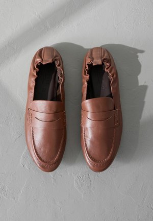Zapatos de piel marrón tipo slip-on con punta redonda, abertura elástica y detalle de costura decorativa en la parte superior. Textura suave.