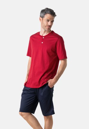 Homme aux cheveux gris portant une chemise rouge à manches courtes avec deux boutons et un short bleu marine, debout les mains dans les poches.