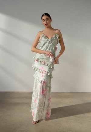 Nainen seisoo kukallisessa, röyhelöisessä maksimekossa, jossa on ohuet hihnat, käsissään sopiva pieni laukku, yksinkertaisen vaalean taustan edessä.