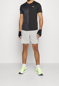 Camisa atlética de manga corta en negro y gris con un patrón de cebra en un lado, combinada con pantalones cortos gris claro y zapatillas de deporte verde neón.
