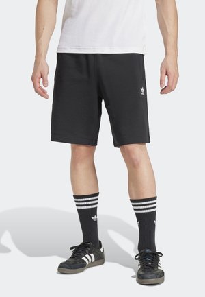 Man draagt zwarte Adidas-shorts, zwarte Adidas-sokken met witte strepen, zwarte Adidas-sneakers en een wit shirt, staand op een witte achtergrond.