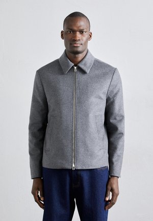 Graue Wolljacke mit spitzem Kragen, Frontreißverschluss und seitlichen Taschen, kombiniert mit dunklen Denim-Hosen. Glatte Textur und taillierte Passform.