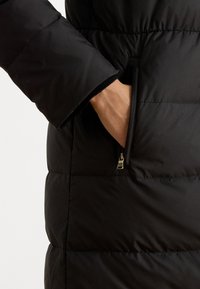 Čierny prešívaný páperový kabát s bočnou vreckom na zips, s mäkkou a hladkou textúrou a priliehavými manžetami na rukávoch.