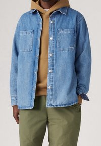 Jasnoniebieska koszula jeansowa z dwoma przednimi kieszeniami, zatrzaskami i casualowym krojem, noszona na brązowej bluzie z kapturem i zielonych spodniach.