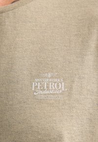 T-shirt en coton beige avec un col rond, arborant un logo graphique blanc portant l'inscription "Motorworks Petrol Industries" ainsi que des accents décoratifs.