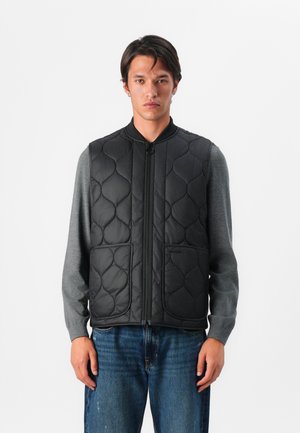 Doudoune noire matelassée avec un col montant, fermeture éclair sur le devant et deux poches à l'avant, portée sur un t-shirt à manches longues gris, avec une coupe décontractée.