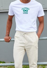 Vit t-shirt med "California League" tryck i grönt, kombinerad med ljusbeige byxor med snörning. Släta texturer, avslappnad passform.