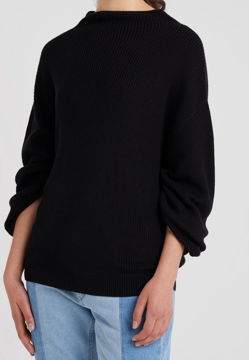 Femme portant un pull en maille côtelée noire avec des manches bouffantes et un jean bicolore bleu clair, debout devant un fond uni.