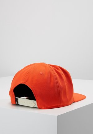 Casquette snapback orange avec sangle en plastique blanche réglable reposant sur une surface blanche contre un fond gris uni.