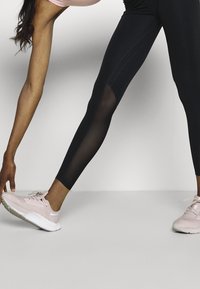 Mustat legginsit, joissa on läpinäkyviä paneeleja, yhdistettynä vaaleanpunaisiin urheilujalkineisiin. Leggingsit ovat sileäteksturoituja ja tiukasti istuvia.