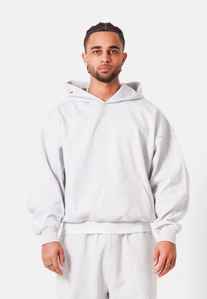 Man met gevlochten haar en baard, gekleed in een lichtgrijze oversized hoodie en bijpassende joggingbroek, staand tegen een witte achtergrond.