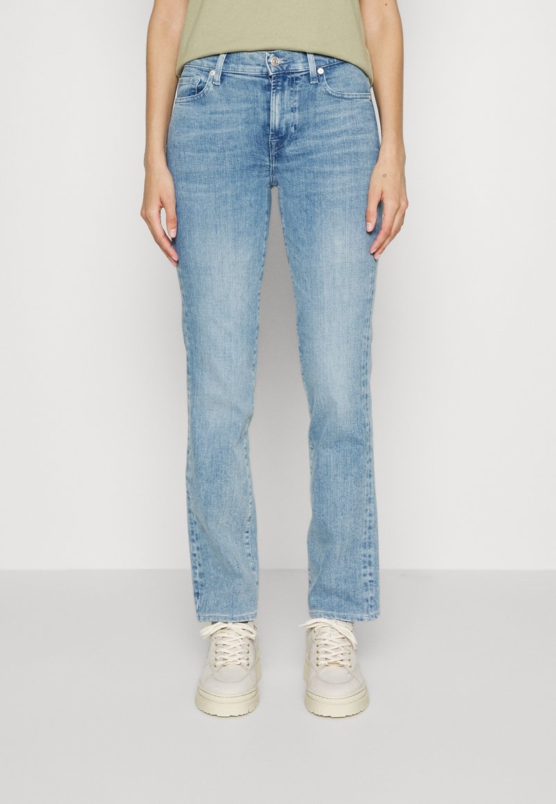 Personne portant un jean droit bleu clair, des baskets beiges et une chemise vert olive, debout devant un fond uni.