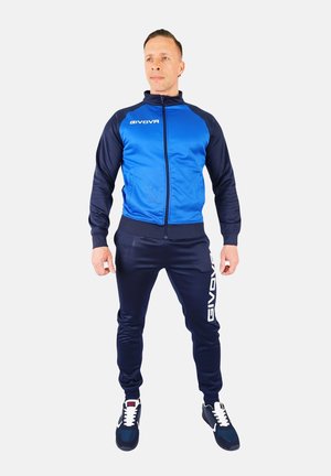 Blue and navy tracksuit with zippered jacket, ribbed cuffs, and pants featuring white logo detail. Worn with sporty shoes.