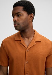 Uomo con capelli ricci corti che indossa una camicia girocollo a maniche corte in un arancione bruciato e un piccolo orecchino a cerchio, che guarda di lato.