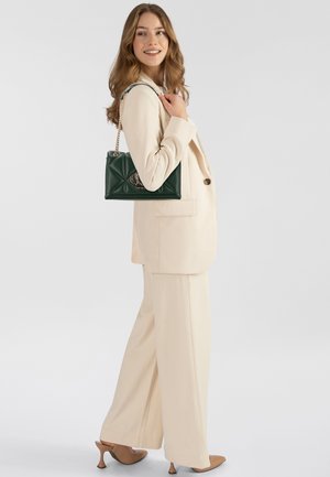 Completo beige su misura con pantaloni larghi, abbinato a una borsa trapuntata verde con hardware dorato e tracolla a catena. Texture liscia e design strutturato.