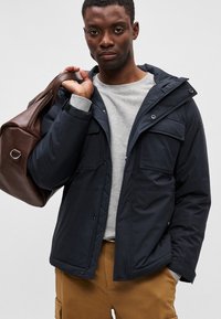 Casaco isolado azul-marinho com capuz, frente abotoada e múltiplos bolsos, combinado com um moletom cinza e uma mala castanha, usado por uma pessoa.