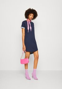 Robe courte marine à manches courtes avec des poignets en denim retroussés, associée à un foulard à motifs roses autour du cou, un sac à main en texture crocodile rose et des bottines à motifs roses.