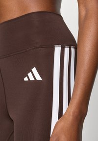 Leggings de sport marron en tissu extensible, avec trois bandes blanches sur le côté et un logo Adidas blanc sur la cuisse.