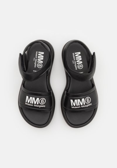 Svarta bärsandaler med justerbart kardborreband, med tryckt vit logotyp "MM6 Maison Margiela" på ovandelen och sulan. Slät textur.