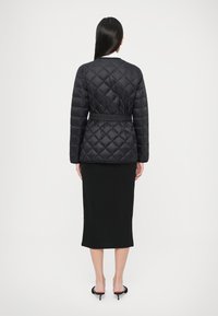 Svart quiltet dunjakke med belte i midjen, med et diamantmønster og rund krage, brukt over et svart tettsittende skjørt.