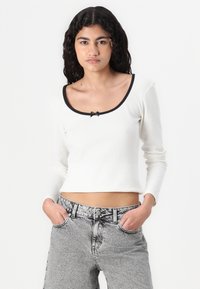Hvit ribbet langermet crop topp med vid halslinning, svart detaljer og en liten sløyfe i sentrum. Kombinert med høy midje lys grå jeans.