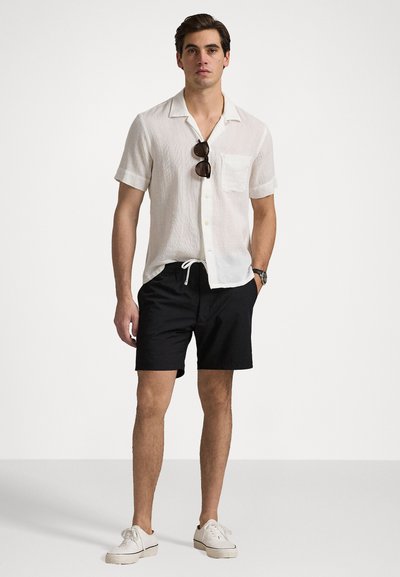 White short-sleeve button-up shirt with a pocket, paired with black drawstring shorts. Worn with white sneakers and sunglasses.