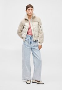 Quiltelt bézs dzseki bolyhos gallérral, alatta rózsaszín ing, laza világoskék farmernadrággal és fekete részletekkel ellátott fehér cipővel párosítva.