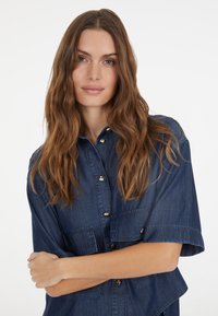 Dark denim shirt with short sleeves, featuring two front pockets, button closure, and a relaxed fit. Smooth texture and uniform color.