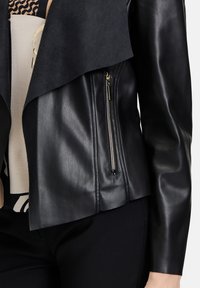 Veste en cuir noir avec un revers en daim, dotée d'une poche zippée sur le côté et d'une texture lisse. Une vue rapprochée met en évidence les détails de conception.