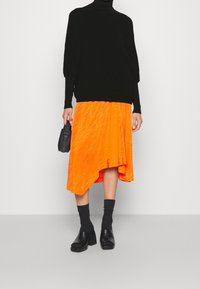 Pull noir oversize à col roulé, jupe asymétrique orange avec finition texturée, chaussures noires à talons blocs et chaussettes montant à la cheville.