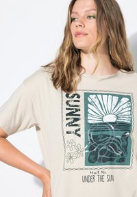 Beżowa koszulka z okrągłym dekoltem i krótkimi rękawami. Ma graficzny nadruk z słońcem, falami oraz napisem "Spotkajmy się pod słońcem" w kolorze czarnym.