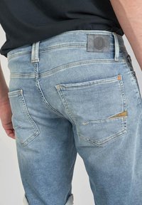 Un jean en denim bleu clair présente une coupe classique, des détails de couture visibles et plusieurs poches, dont une au design angulaire distinctif.