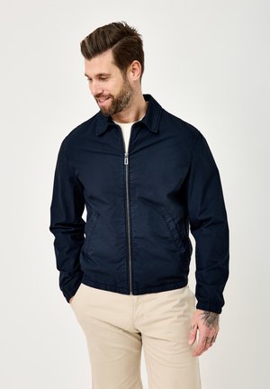 Man with beard wearing navy blue zip-up jacket and beige pants, hands in pockets, looking down and smiling against plain background.
