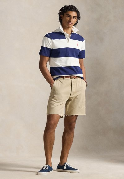 Πόλο πουκάμισο με ναυτικές και λευκές ρίγες, μπεζ σορτς με ξεφτισμένο τελείωμα και ναυτικά αθλητικά παπούτσια. Διαθέτει γιακά και υφασμένη ζώνη.