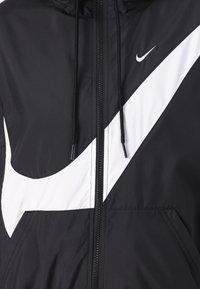 Svartvit Nike vindjacka med en distinkt stor swoosh-design, dragkedja, justerbar dragsko i huvan och slät syntetisk tyg.