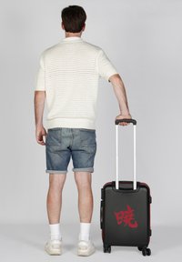 Maleta dura negra con acentos rojos y diseño de texto, equipada con un mango retráctil y cuatro ruedas, llevada por una persona vestida con ropa casual.