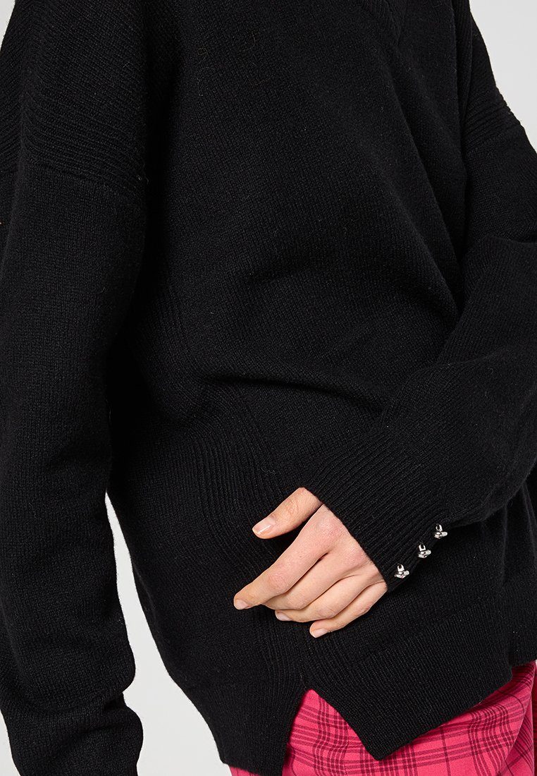 Maglione nero a maglia con vestibilità ampia, caratterizzato da una texture a coste, spacchi laterali e dettagli in metallo decorativi sui polsini delle maniche.