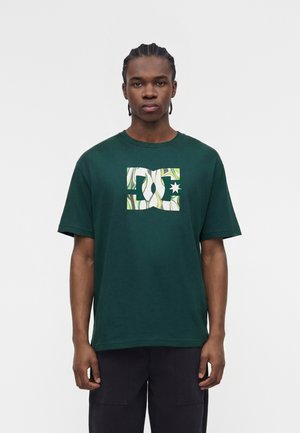 Νεαρός άνδρας με πλεξούδες που φοράει σκούρο πράσινο μπλουζάκι με λευκό και πράσινο μοτίβο λογότυπο DC στο στήθος, στέκεται μπροστά σε απλό φόντο.