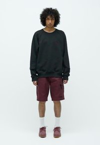 Hombre joven con cabello rizado, vistiendo una sudadera negra de manga larga, pantalones cortos tipo cargo burdeos, calcetines blancos y zapatos burdeos, de pie contra un fondo blanco.